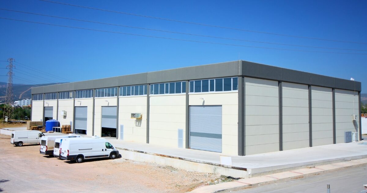 A large, modern industrial warehouse building with cream-coloured walls, grey columns, and several large roller-shutter doors