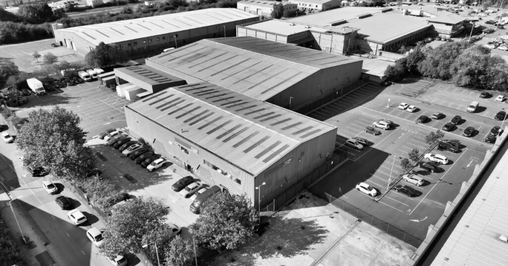 A black and white aerial view of large industrial warehouses surrounded by outdoor parking lots filled with cars.