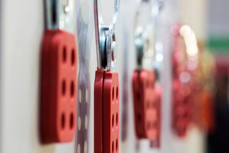 Close-up view of red lockout tagout hasps hanging in a row against a white wall.